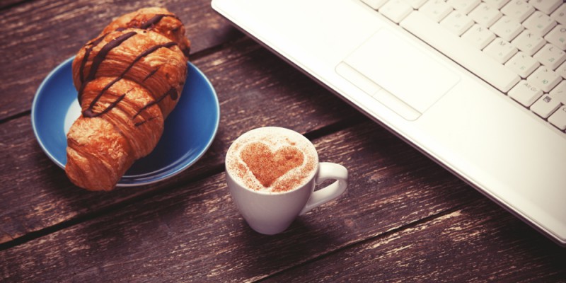 Croissant and cup of coffee with laptop on wooden table.